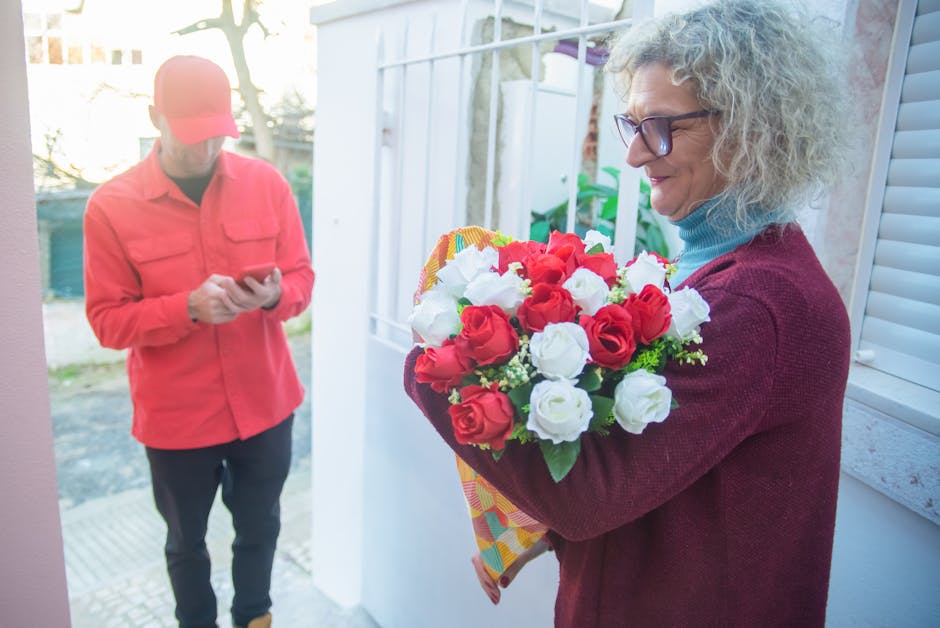 A woman with curly gray hair and glasses holding a vibrant bouquet of fresh red and white roses wrapped in decorative paper, poised near a doorway during daytime. The bouquet features lush green leave