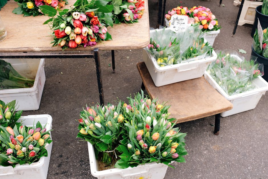 At a flower market, several white plastic crates and small wooden tables display vibrant floral arrangements and bouquets of fresh tulips in shades of pink, yellow, and white, with lush green foliage.