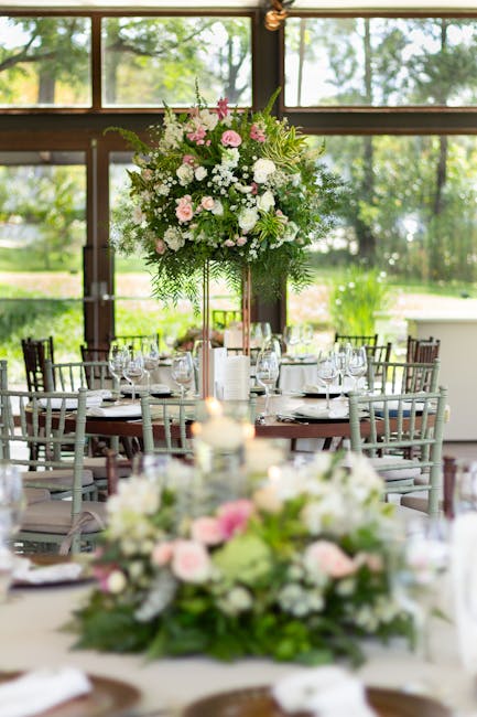 A large, lush floral arrangement featuring pink and white roses, white hydrangeas, and greenery, elevated on a tall stand in the center of a well-lit dining area with large windows showing a garden vi