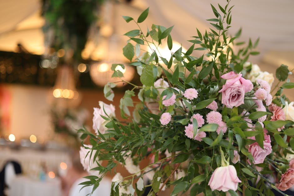 A floral arrangement featuring a lush mixture of pink roses, small light pink daisy-like flowers, and abundant green foliage, including long, slender leaves and smaller rounded leaves, arranged in a n
