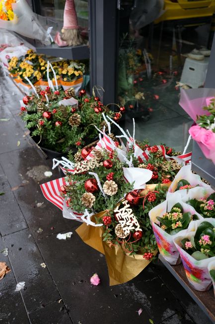 A festive floral arrangement featuring a bouquet of fresh evergreen branches decorated with shiny red baubles, pinecones, and sprigs of holly berries, wrapped in white and red striped paper with a 'Me