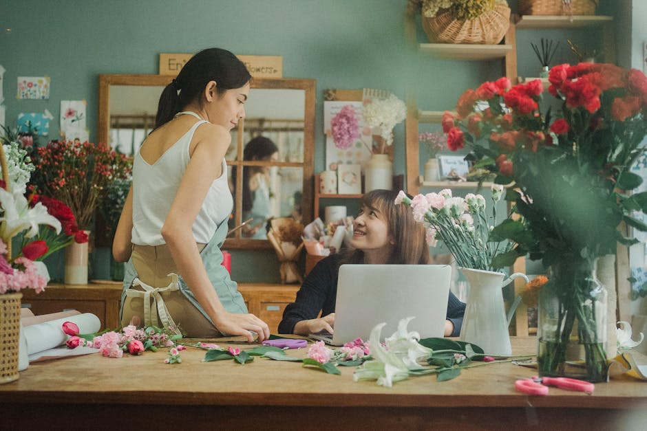 A florist and a customer inside a flower shop engaging in a conversation about a floral gift. The florist, wearing a white sleeveless top and beige apron, stands on the left side of the image, leaning