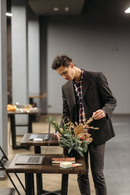 A young man with curly hair dressed in a dark pinstripe blazer and checked shirt arranging a floral bouquet on a wooden table in an indoor setting. The bouquet features a mix of fresh flowers includin