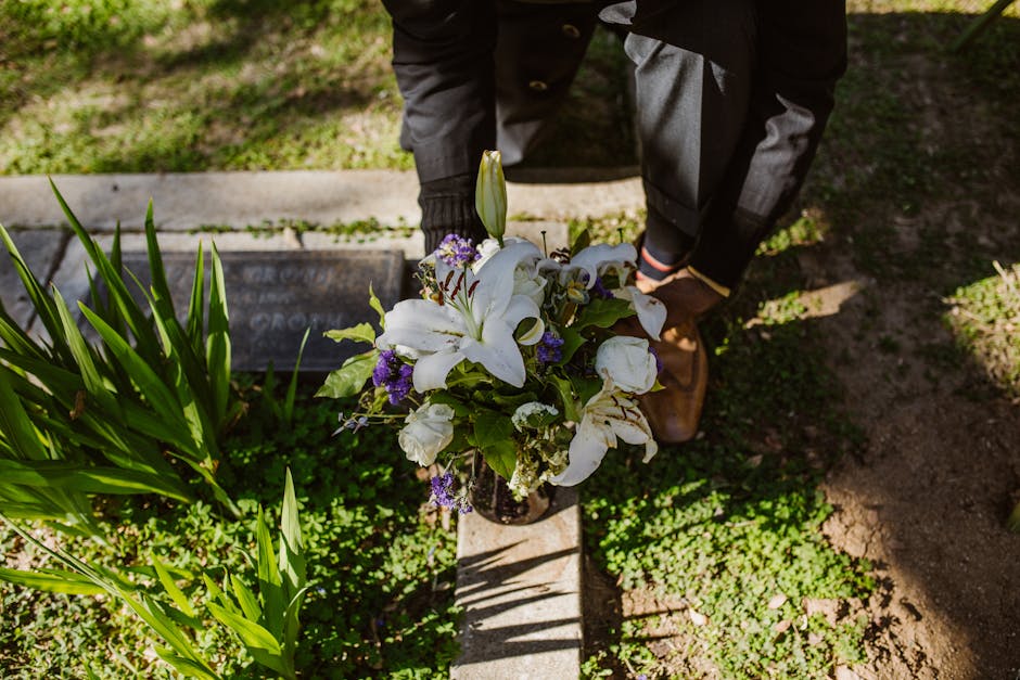 A floral arrangement featuring white lilies with large, open petals and prominent stamens, complemented by smaller purple flowers and green foliage, placed in a glass vase. The bouquet is held by a pe