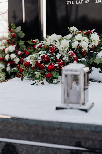A floral arrangement placed on a white table surface, consisting of predominantly white chrysanthemums and lush green foliage, complemented by deep red roses and smaller red berries for contrast. The