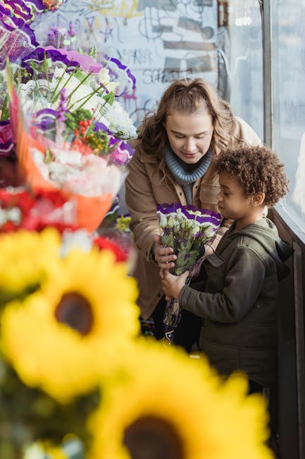 A young woman and a small child are inside a flower shop near a large window, engaging in a floral gift exchange. The woman, with wavy blonde hair and dressed in a beige coat and a blue turtleneck, is