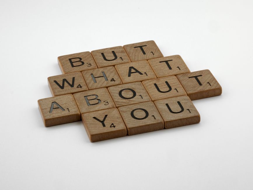 A close-up of wooden Scrabble tiles arranged on a white background spelling out 'BUT WHAT ABOUT YOU.' The tiles have black letters and are slightly textured, with some tiles showing small numbers indi